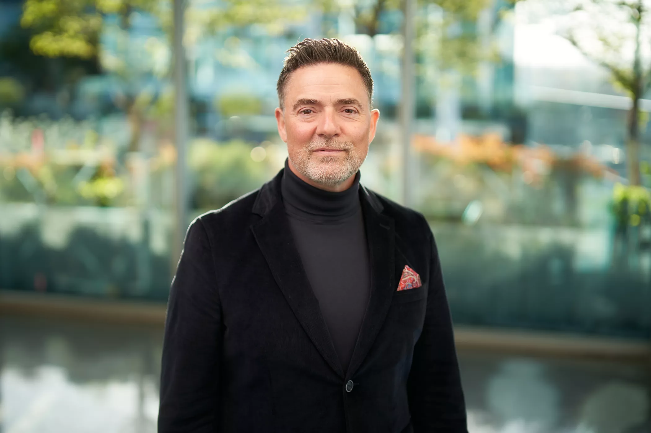 A man wearing a black blazer and a dark turtleneck stands in a modern outdoor setting with glass walls and greenery in the background. The outfit is accessorized with a red patterned pocket square. The scene is brightly lit, emphasizing a professional and stylish appearance.